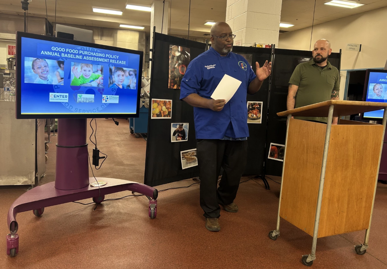 Two men standing in a school district kitchen facility next to a monitor. One man is wearing a blue chef coat while speaking.
