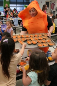 202504LakeTravisISDCarrotDay A school cafeteria worker dressed in a carrot costume serves carrots to school children