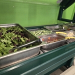 A school chef pours a measuring cup of liquid over a silver tray of scratch cooked food