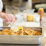 2025ChefAnnGetSchoolsCookingpouring-1236-x-850 A school chef pours a measuring cup of liquid over a silver tray of scratch cooked food
