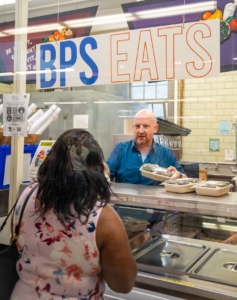 A man serves food in a school cafeteria under a banner that reads BPS EATS