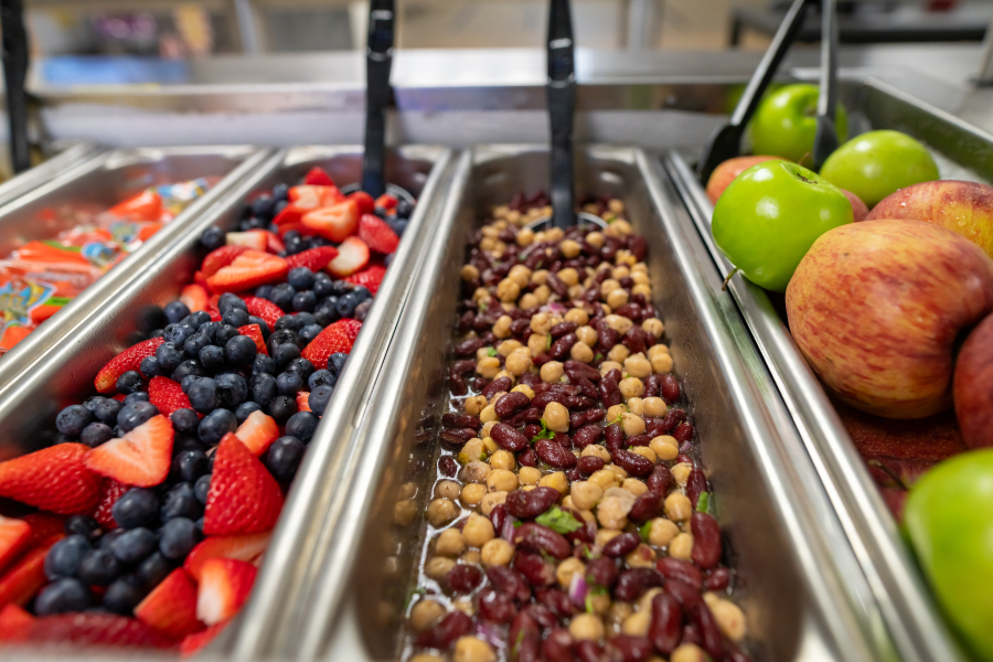 Serving containers of cut fruit, bean salad, and whole apples in a cafeteria lunch line