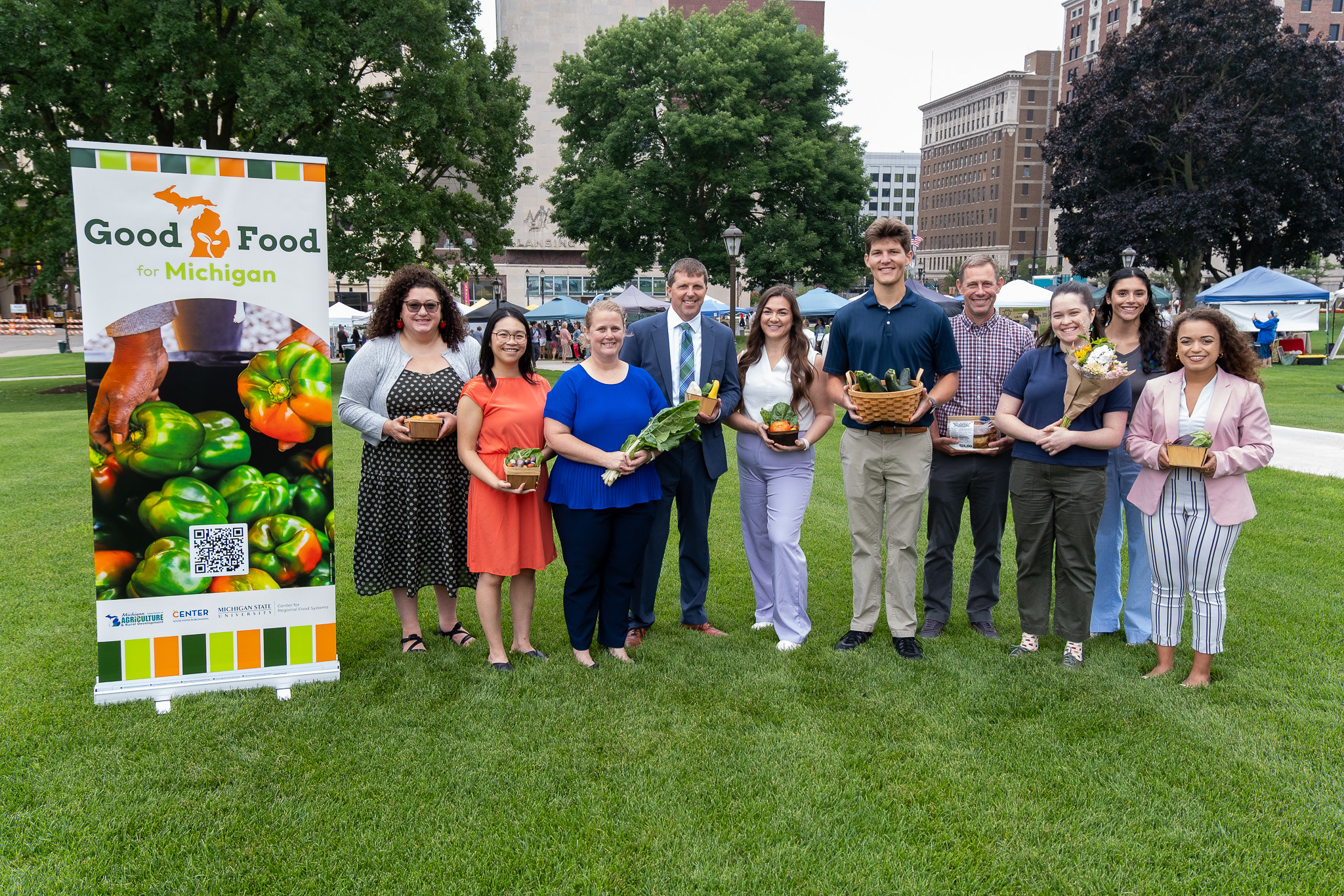 Food systems leaders hold Michigan-grown vegetables and flowers standing beside a Good Food for Michigan banner on the lawn of the Michigan Capitol Farmers Market
