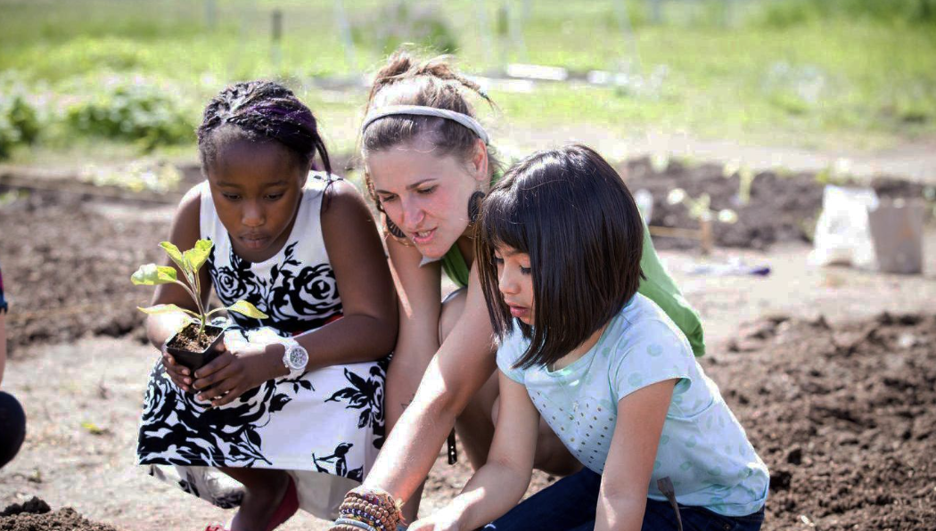 An adult and two children tend to a dirt field. one child holds a potted plant.